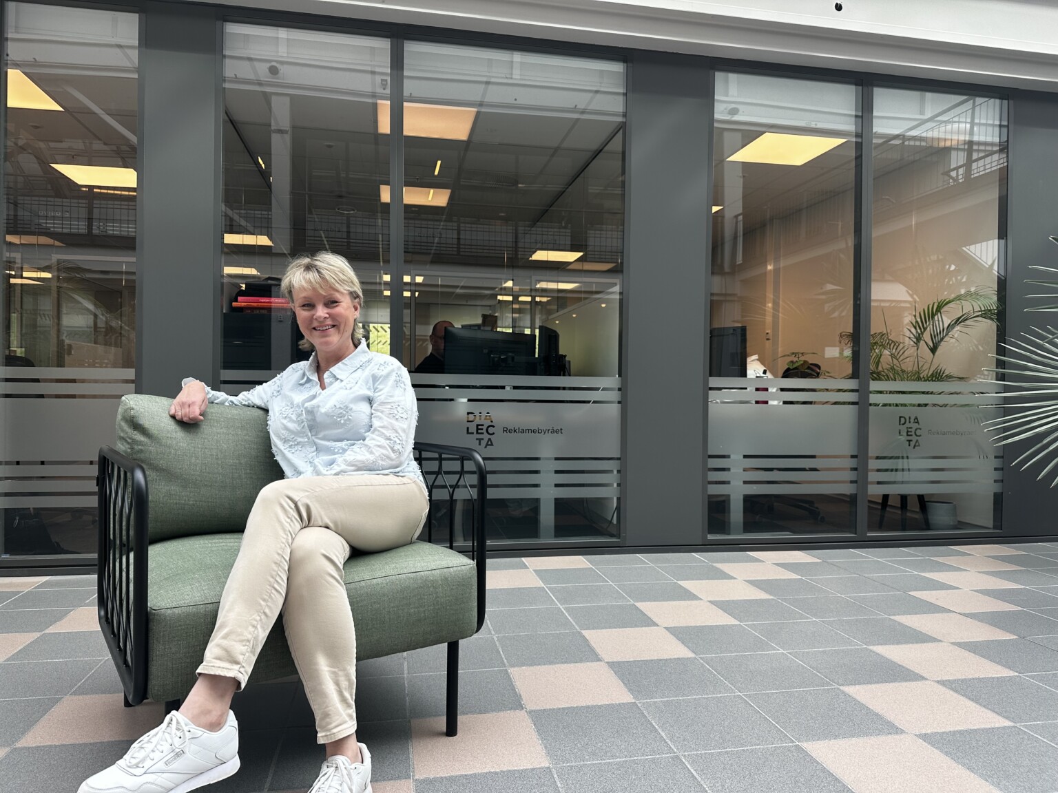 Smilende kvinne i en grønn lenestol foran et glassparti med Dialecta-dekor