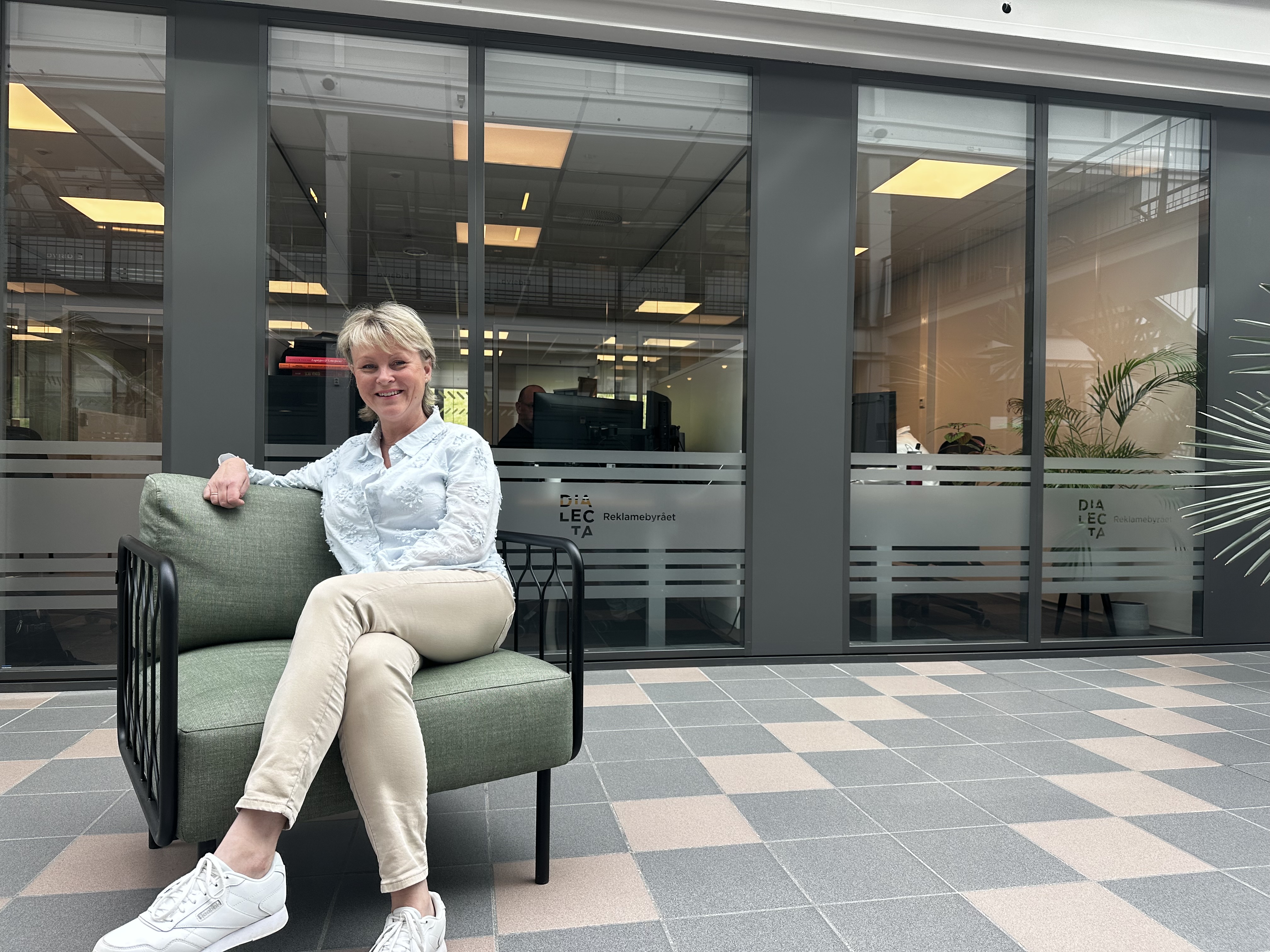 Smilende kvinne i en grønn lenestol foran et glassparti med Dialecta-dekor