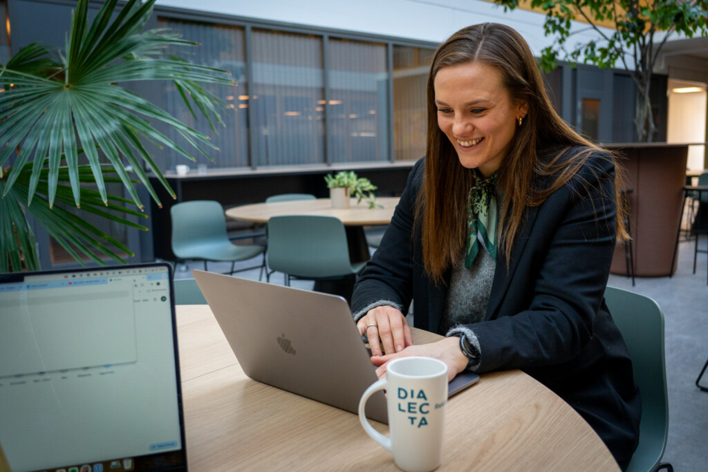 Smilende kvinne jobber på bærbar PC med Dialecta-krus på bordet
